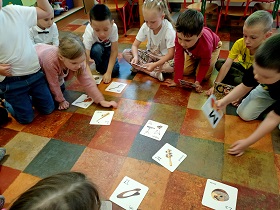 Dzieci siedzą na podłodze i trzymają w dłoniach kartoniki z narysowanymi instrumentami. 