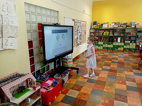 Dziewczynka w sukience stoi przy monitorze, na którym wyświetlony jest slajd z wirtualnymi kostkami. 