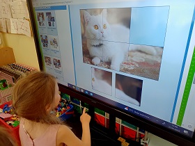 Dziewczynka stoi przy monitorze, na którym wyświetlone jest zdjęcie z białym kotem. Zdjęcie podzielone jest na sześć małych kwadratów. Jednego elementu brakuje. Dziewczynka wybiera element. 