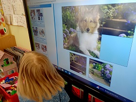 Dziewczynka stoi przy monitorze, na którym wyświetlone jest zdjęcie z psem. Zdjęcie podzielone jest na sześć małych kwadratów. Jednego elementu brakuje. Dziewczynka wybiera element. 