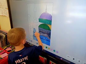 Chłopiec stoi przy monitorze i maluje palcem obraz. Sam dobiera kolory, których używa. 