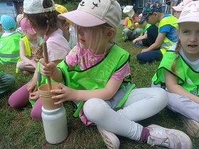 Dzieci siedzą na trawie, jedna dziewczynka trzyma maselnicę – urządzenie do ręcznego wyrabiania masła.