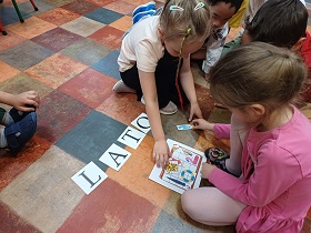 Dwie dziewczynki układają wakacyjne przedmioty w walizce. Walizka oraz ów przedmioty są wydrukowane. Powyżej z drukowanych liter ułożony jest wyraz LATO.
