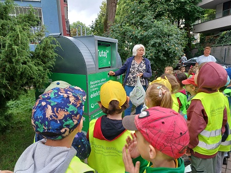 Dzieci w odblaskowych kamizelkach stoją przed zielonym pojemnikiem na elektrośmieci. Pani Hania wrzuca do niego uzbierane baterie. 