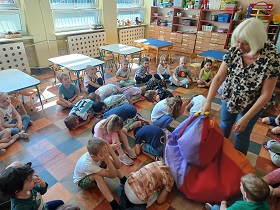Dzieci siedzą na podłodze w kole. Część z nich jest w środku kuląc się i chowając. Pani Hania przykrywa jedno z dzieci chustą animacyjną. 
