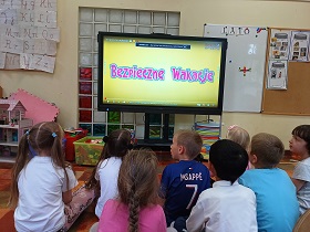 Dzieci siedzą na podłodze przed monitorem edukacyjnym. Na ekranie wyświetla się na żółtym tle ,,Bezpieczne wakacje”.