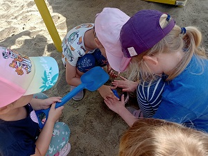 Dzieci kucają na piasku. Jedna dziewczynka trzyma w dłoniach niebieską łopatkę. 