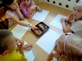 Dzieci siedzą przy stoliku i rysują kredkami na białych kartkach. Na środku stolika leży pudełko z kredkami. 