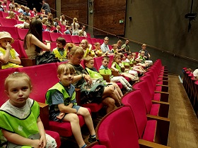 Dzieci w odblaskowych kamizelkach wraz z paniami siedzą na fotelach w sali teatralnej. 