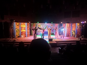 Na scenie widać czterech aktorów, którzy wznoszą ręce do góry. Za nimi stoi kolorowa dekoracja. 