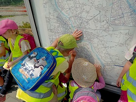 Dzieci stoją przy mapie na przystanku autobusowym. Dzieci mają na sobie kamizelki odblaskowe. 