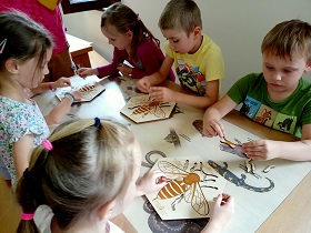 Dzieci siedzą przy stoliku parami, na przeciwko siebie i układają drewniane puzzle przedstawiające budowę pszczoły. 