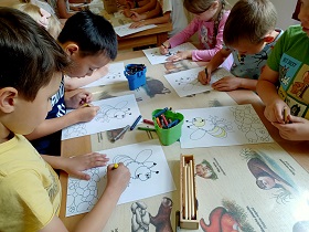 Dzieci siedzą przy stoliku i kolorują kolorowankę pszczoły. Na stoliku znajdują się pudełka z kredkami. 