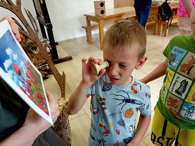 Chłopiec patrzy przez przyrząd, który pokazuje mu jak widzi pszczoła. Chłopiec przykłada przyrząd do oka i spogląda na obrazek przedstawiający łąkę. 
