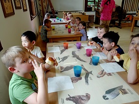 Dzieci siedzą przy trzech stolikach i jedzą bułki. Przed nimi leżą chusteczki oraz stoją kubeczki z herbatą. 