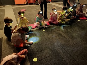 Dzieci siedzą na podłodze na kolorowych poduszkach. Mają na czołach latarki czołówki i spoglądają na zielone kółka, świecąc na nie. 