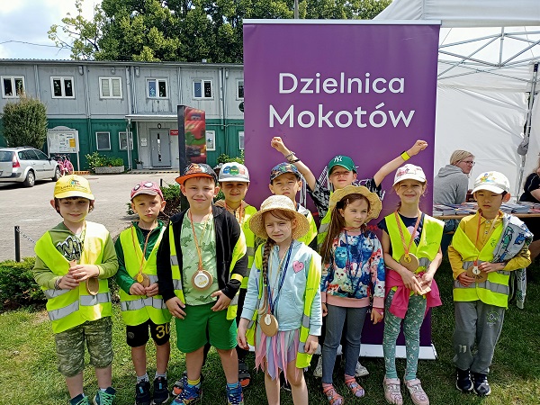 Dzieci w odblaskowych kamizelkach stoją na tle napisu Dzielnica Mokotów i pozują do zdjęcia. 