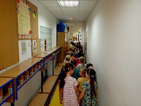 Dzieci stoją w parach w korytarzu. Obok nich stoją szafki i ławki.