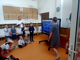 Pani w szarej sukience stoi przy monitorze i odczytuje pytanie z granatowej planszy. Wokół monitora stoją i siedzą dzieci. 