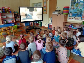 Dzieci siedzą na podłodze przed monitorem, na którym wyświetlany jest film, gdzie dziewczynka układa swojego tatę w pozycji bezpiecznej. 