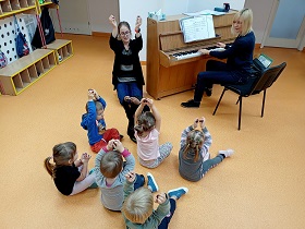 Dzieci siedzą na podłodze razem z panią. Wszyscy trzymają w dłoniach kasztany. Pani pokazuje dzieciom jak nimi grać. Obok dzieci i pani gra na pianinie pani.