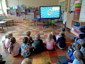 Dzieci siedzą na podłodze przed monitorem i oglądają filmik o służbach ratunkowych – na filmie wyświetla się numer alarmowy 999.