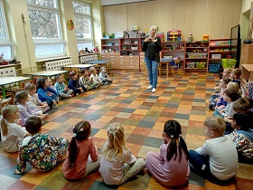 Dzieci siedzą w półkolu na podłodze. W środku półkola stoi pani i wskazuje na siebie. 