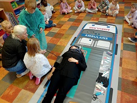 Pani w czarnym ubraniu i szaliku na głowie leży na noszach, na planszy ambulansu. Dwoje dzieci wraz z panią mierzy jej ciśnienie. 