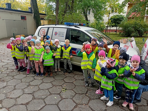 Grupa dzieci w kamizelkach odblaskowych stoi przy samochodzie ratownictwa medycznego i pozuje do zdjęcia. 