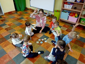 Grupa dzieci i pani siedzą na podłodze. Pani oraz kilkoro dzieci zgłasza się. Przed dziećmi i panią leżą obrazki.