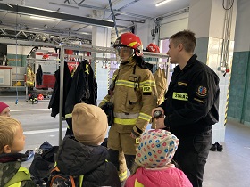 Przed dziećmi stoi dwóch strażaków. Po lewej stronie stoi strażak w stroju do akcji, na głowie ma założony kask. Obok niego stoi mężczyzna w czarnym mundurze z napisem straż.