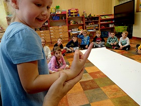 Dzieci siedzą w siadzie skrzyżnym na podłodze i obserwują dziewczynkę, która spogląda na swój palec, który jest umazany pieprzem. 