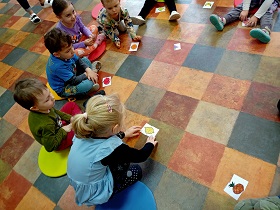 Dzieci siedzą w kole na podłodze. Dziewczynka w niebieskiej sukience łączy obrazek cytryny w całość. Przed pozostałymi dziećmi również leżą obrazki owoców. 