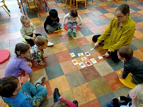 Dzieci siedzą w kole na podłodze wraz z panią. Pani łączy dwie połówki obrazka w całość. 