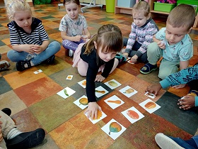 Dzieci siedzą w kółku. Przed nimi leżą obrazki dyń. Dziewczynka w czarnej bluzce sięga po jeden z obrazków. Reszta dzieci się temu przygląda. 