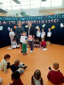 Na zdjęciu widać chłopca z dyplomem i nagrodą w rękach oraz dwie panie, które stoją za nim. Za nimi stoi granatowy parawan z napisem: konkurs świąteczne puzzle – święto niepodległości, a także z papierowymi puzzlami. 