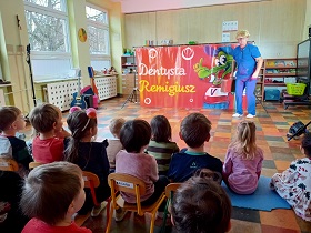 Dzieci siedzą jako publiczność na przedstawieniu. Przed nimi występuję aktor przebrany za dentystę, w blond peruce. Obok niego znajduje się dekoracja sceniczna z napisem Dentysta Remigiusz. 