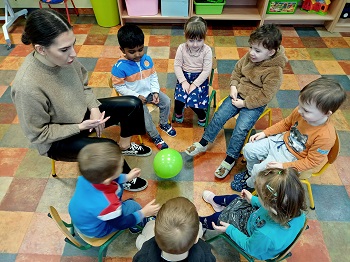 Dzieci siedzą w kółku na krzesełkach ustawionych blisko siebie. Próbują odbijać zielony balon, tak by nie spadł na podłogę.