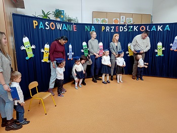 Dzieci wraz z rodzicami stoją obok siebie. Za nimi widać granatowy parawan z napisem pasowanie na przedszkolaka oraz papierowe kredki. 