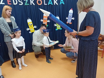 Pani kuca przy dziecku i wręcza mu książkę z dyplomem. Za dzieckiem stoi tata. Obok nich stoi mama z dziewczynką oraz pani z papierowym ołówkiem w ręku. 