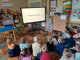 Dzieci siedzą na podłodze przed monitorem, na którym wyświetlany jest film. Napis na ekranie: Czym jest wolontariat?