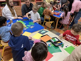 Przy stole z kolorowymi papierami siedzą rodzice i dzieci, które wycinają różne kształty.