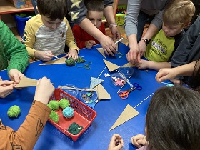 Dzieci siedzą i szpatułkami smarują trójkąty. Na stole ustawiony jest koszyk z włóczką oraz kolorowe elementy do ozdobienia choinki.