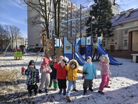 Siedmioro dzieci stoi na środku placu zabaw i macha rączkami. Wokół jest śnieg.