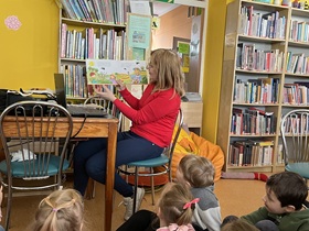 Pani siedzi na krzesełku, w dłoniach trzyma książkę z rysunkami, dotyczącymi oznak wiosny. Dzieci siedzą i patrzą na ilustracje.