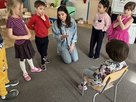 Na krzesełku siedzi dziewczynka, przed nią stoją w półkolu dzieci oraz klęczy pani.