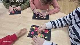 Troje dzieci siedzi przy stoliku i wykonuje pracę plastyczną pn. "wydrapywanka" o tematyce kosmosu.
