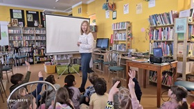 Kilkoro dzieci siedzi na podłodze, czworo z nich ma podniesione rękę do góry, przed nimi stoi kobieta, za kobietą jest ustawiony ekran projekcyjny, w tle widać wyposażenie biblioteki.