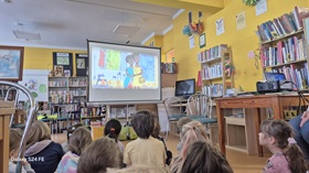 Kilkoro dzieci siedzi na podłodze i ogląda film edukacyjny na ekranie projekcyjnym "Droga po wodę w Afryce", w tle widać wyposażenie biblioteki.