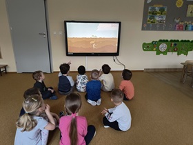 10 dzieci siedzi na podłodze w sali. Odwrócone są w kierunku monitora, na którym wyświetlony jest film o rozwoju roślin.
